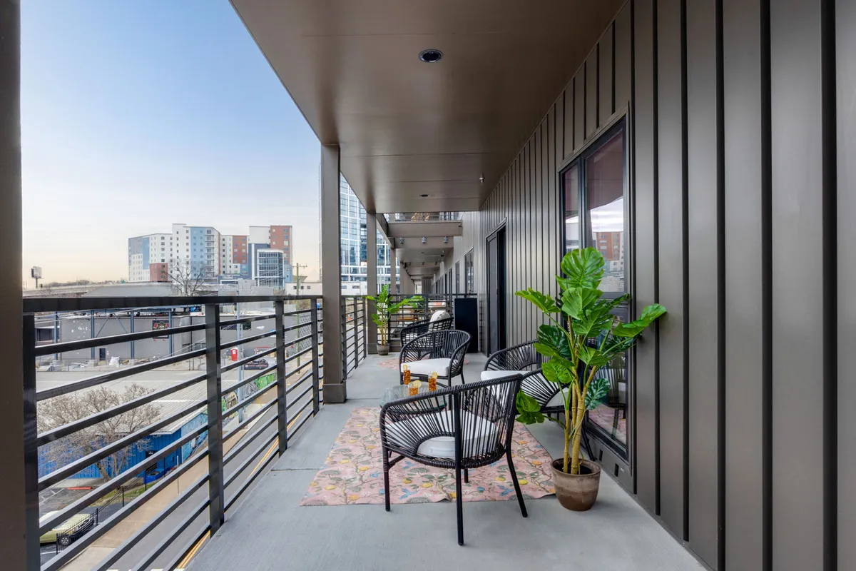 A cozy balcony seating area with potted plants, a decorative rug, and a partial city view. The setup invites relaxation, morning coffee, or evening chats while overlooking the cityscape.
