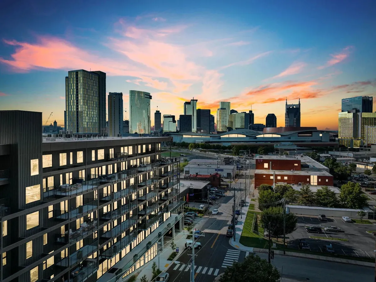 A striking view of a modern apartment building set against the Nashville city skyline at sunset. The vibrant sky and tall buildings evoke the city's energy and legendary musical atmosphere. Pedestrians and nearby cafes hint at urban exploration and nightlife.
