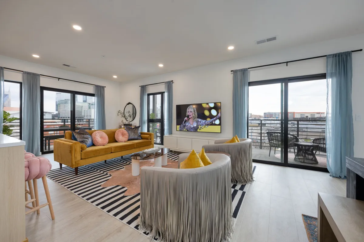 The image showcases a vibrant, Dolly Parton-inspired living room featuring a bold yellow sofa, whimsical fringed chairs, and a large TV. Sliding doors open to a private balcony with sunset city views, creating a welcoming space for socializing, relaxing, or enjoying a movie night in Nashville.