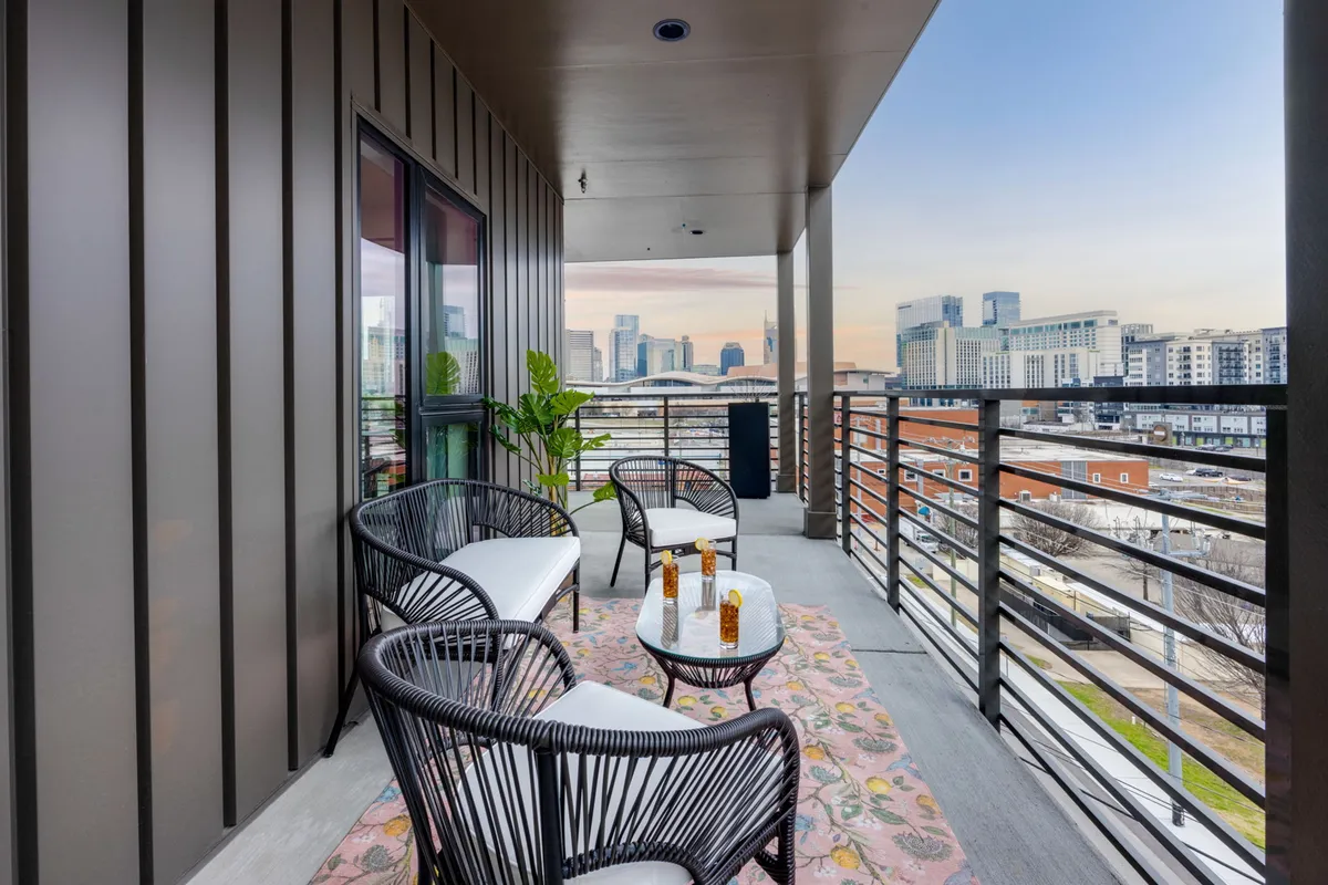 A stylish balcony with comfortable seating, a decorative rug, and a stunning view of the Nashville city skyline at sunset. The mood is inviting, ideal for socializing or capturing Instagram-worthy skyline photos.
