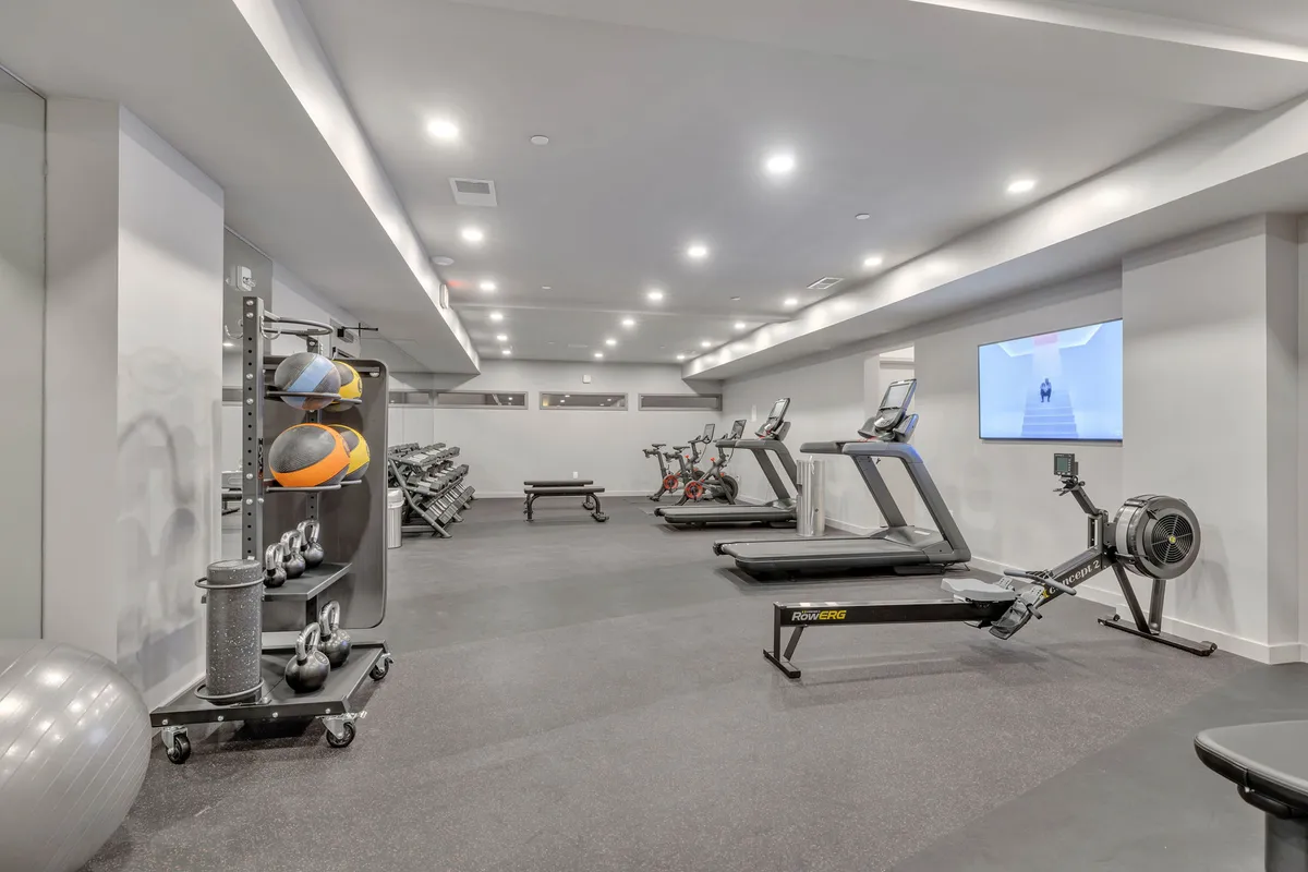 A bright, modern gym with multiple treadmills, exercise bikes, rowing machines, dumbbells, and medicine balls. The space is energetic and well-lit, designed for both solo and group workouts.