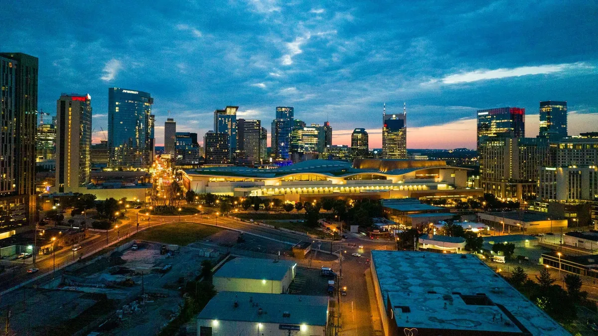 A panoramic view of the city skyline at dusk, with illuminated buildings, a cloudy sky, and a main roadway leading into the heart of downtown. The image exudes energy, sophistication, and the promise of vibrant city nights.