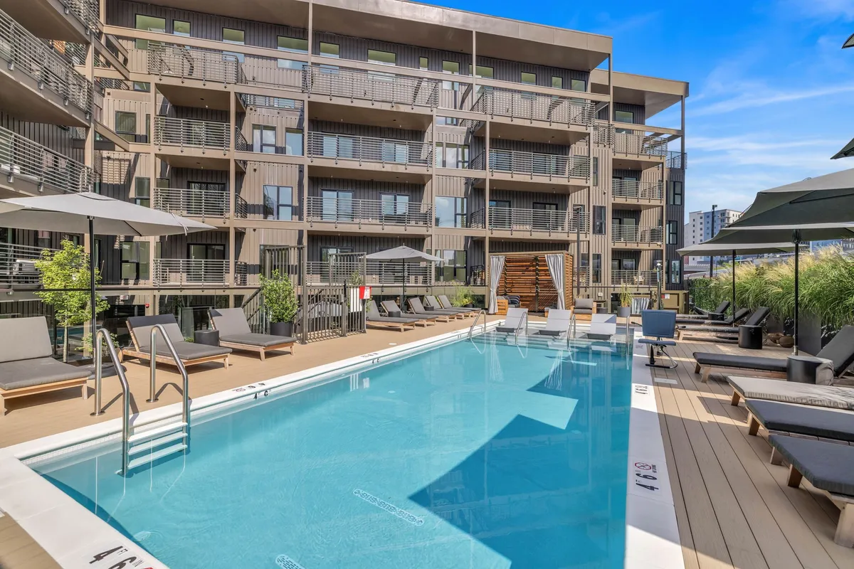 A bright and inviting exterior view of the building's swimming pool area, featuring deck chairs, umbrellas, and the building facade in the background. The scene captures a relaxed atmosphere perfect for sunny days spent swimming, sunbathing, and socializing.