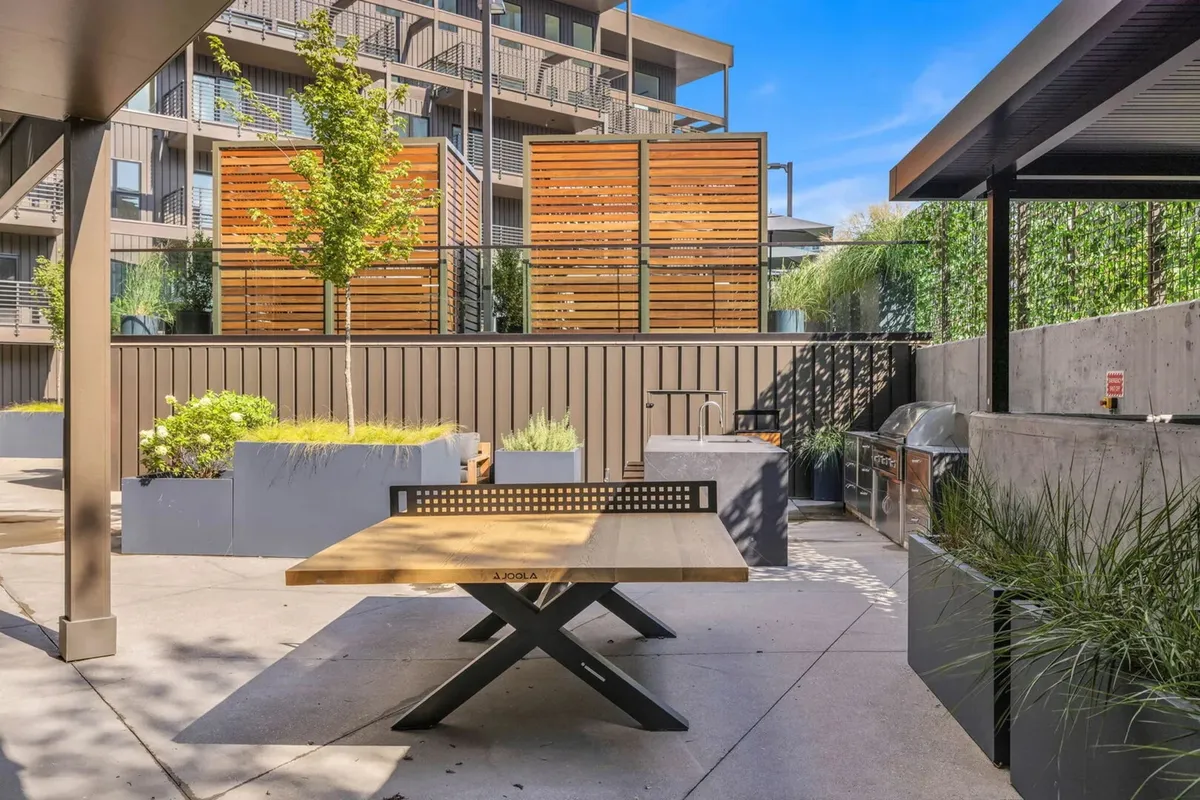 A welcoming outdoor lounge area featuring a wooden picnic table, decorative wooden screens for privacy, greenery in planters, and a designated grill area. The space is ideal for BBQs, casual gatherings, and enjoying meals under open skies.