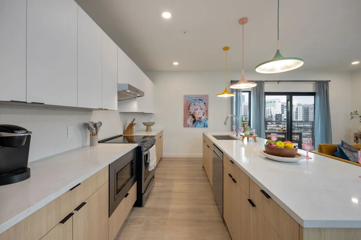 A contemporary kitchen scene with a spacious island, bright pendant lights, and a fruit bowl as the centerpiece. Wall art featuring a woman and a window with a city view add personality and brightness, while bar seating ensures the chef is never left out of the conversation.